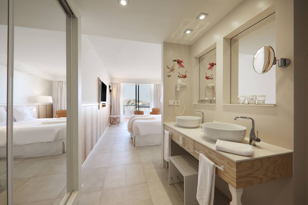Um banheiro de hotel moderno com uma pia grande, espelho e duas camas visíveis ao fundo.