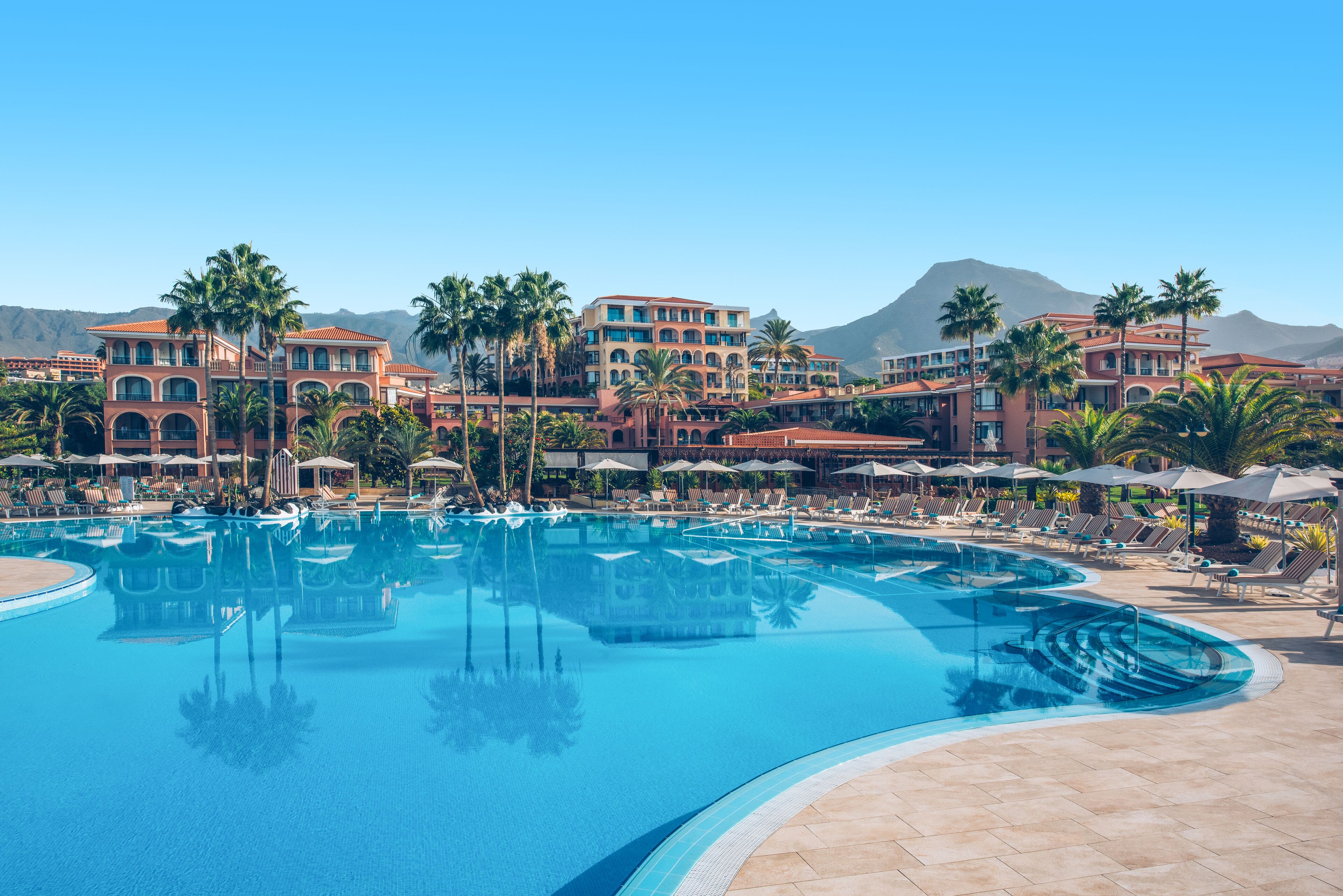 A large swimming pool with lounge chairs and umbrellas, surrounded by palm trees and a hotel building with multiple balconies.