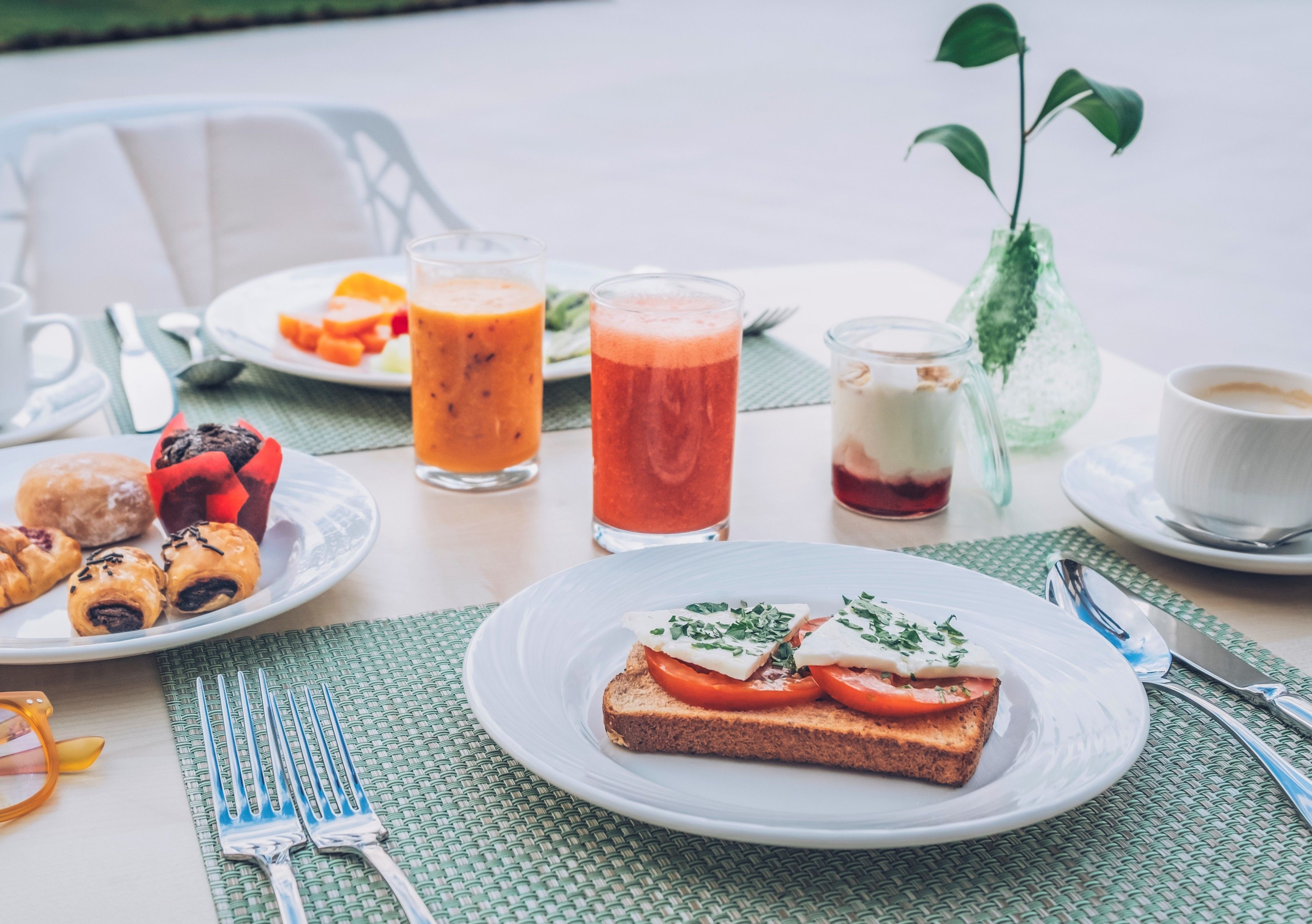 Un desayuno con tostadas, un vaso de zumo de naranja, un vaso de zumo rojo, una taza de café y un pequeño florero con una planta.