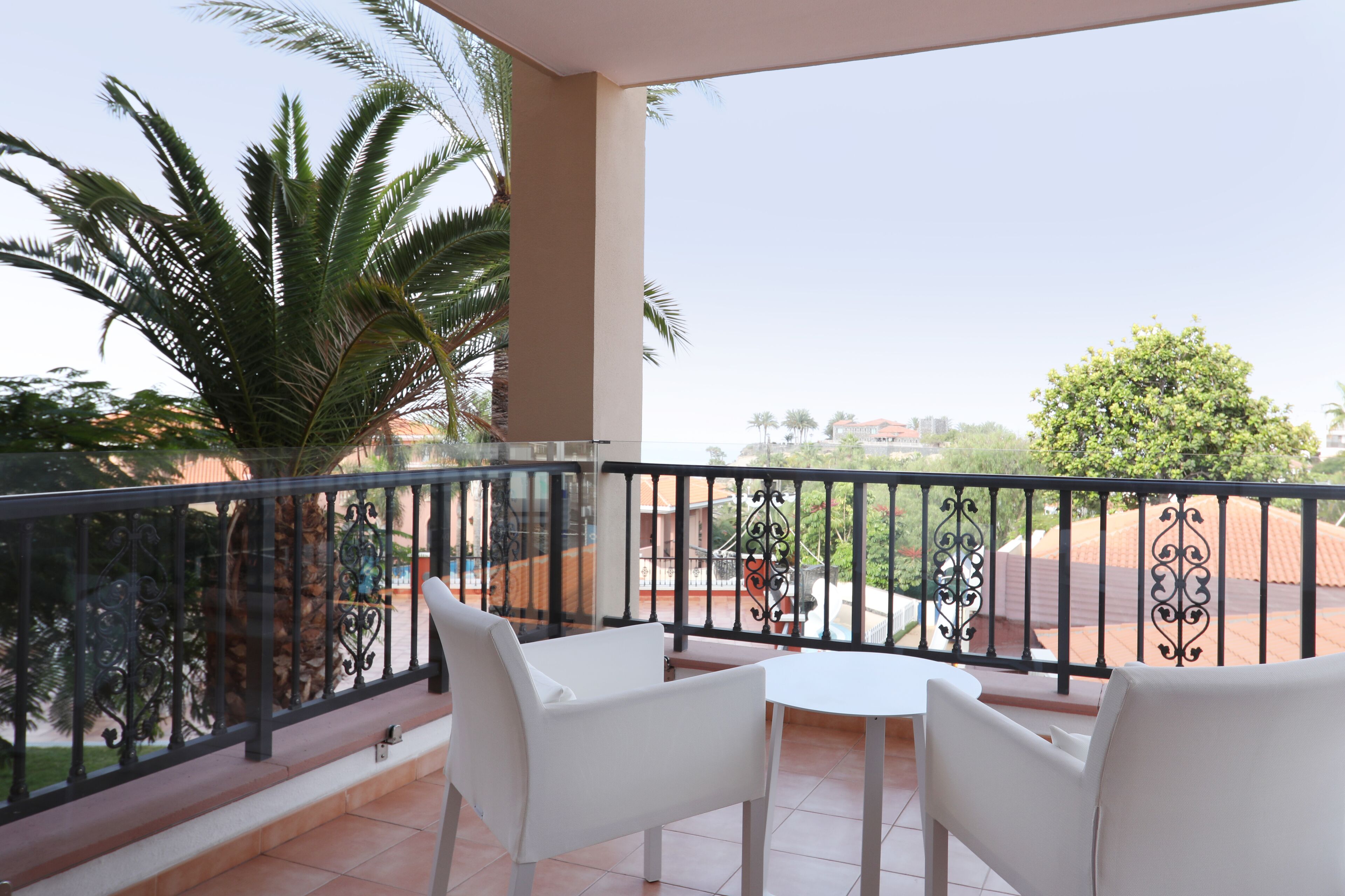 Een balkon met witte stoelen en een tafel, een palmboom en uitzicht op gebouwen en bomen.