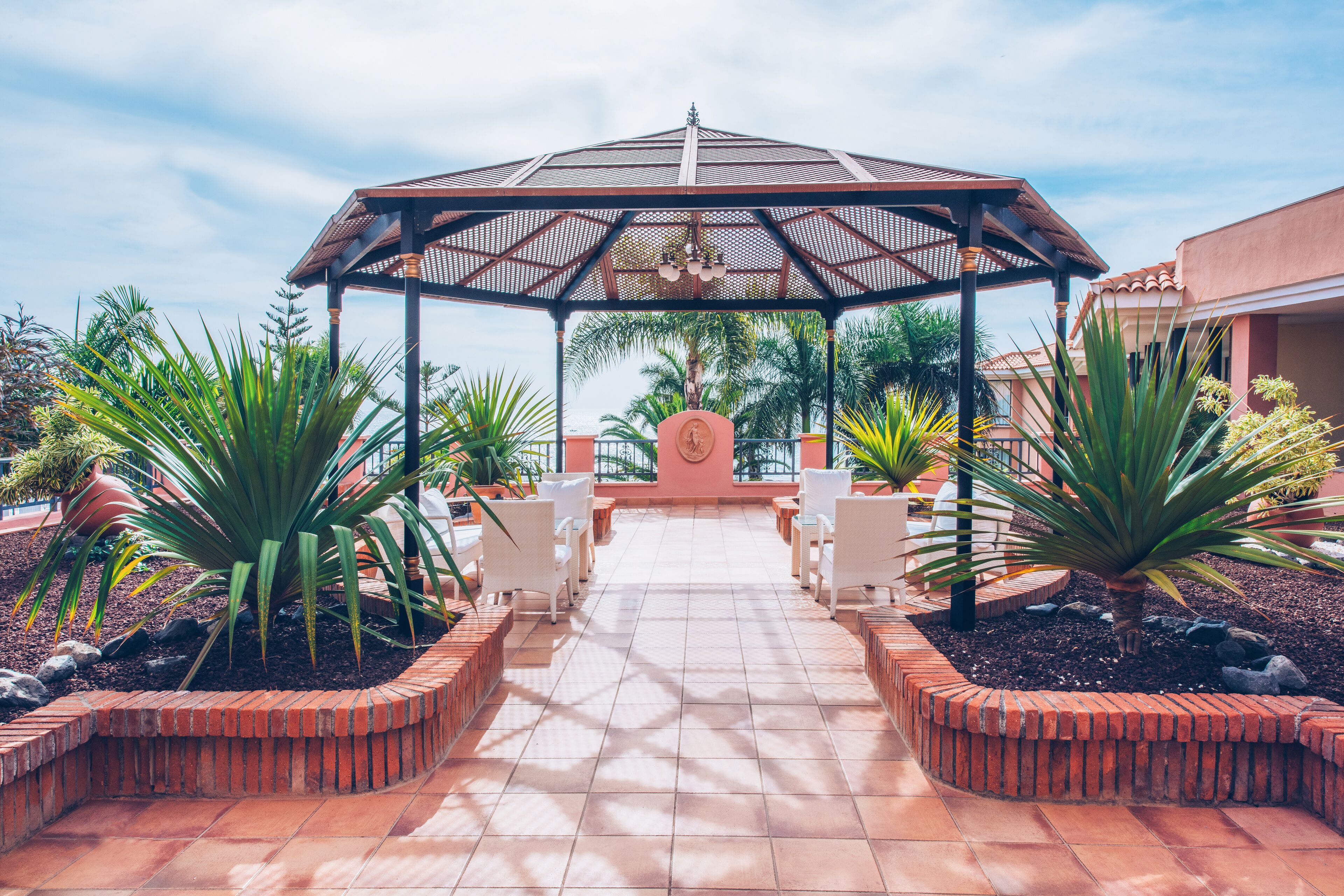 Ein überdachter Patio-Bereich mit gefliestem Boden, umgeben von Ziegelkübeln und weißen Stühlen.