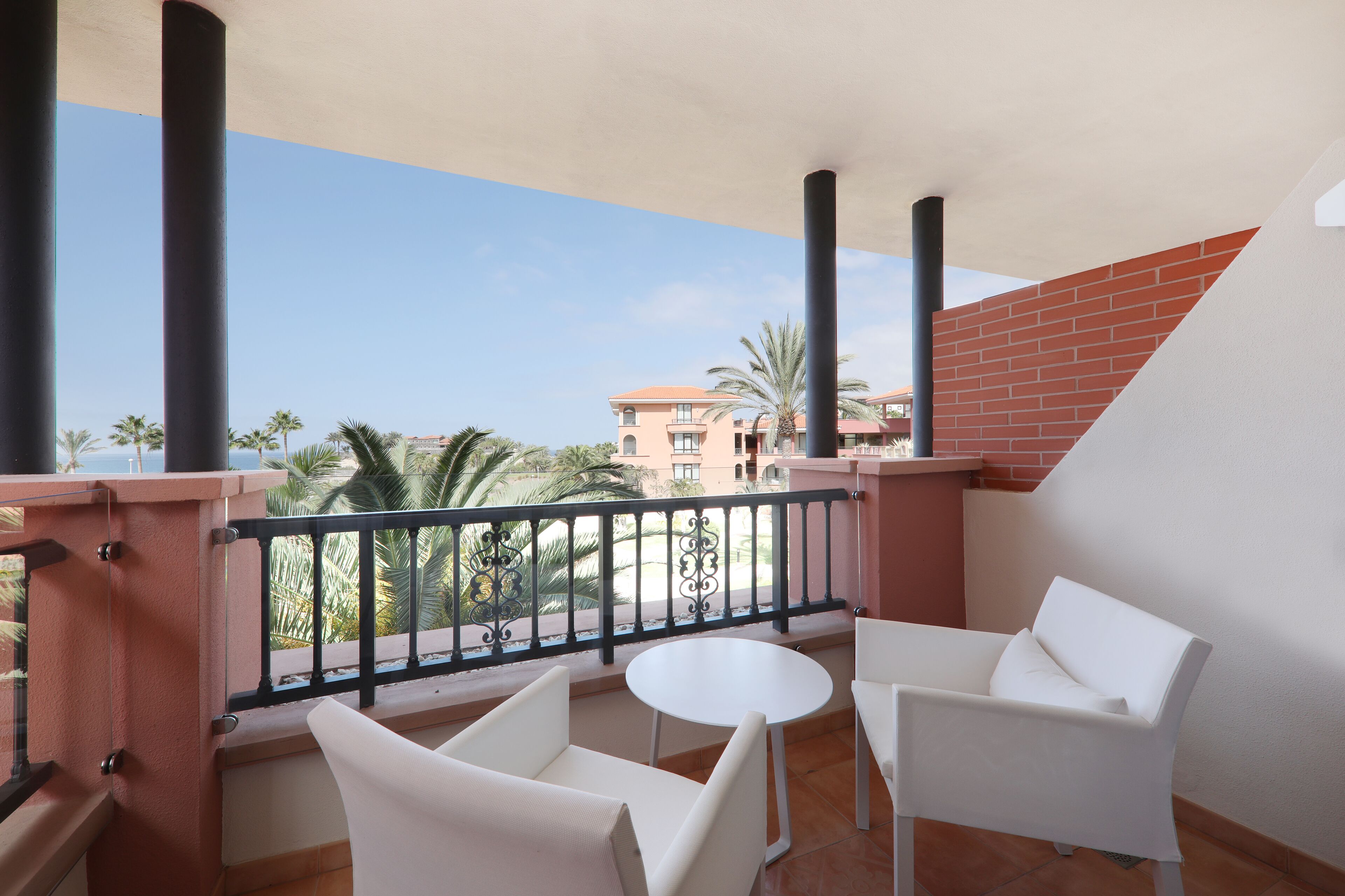 A balcony with white chairs and a small round table, overlooking a building with a red brick wall and palm trees.