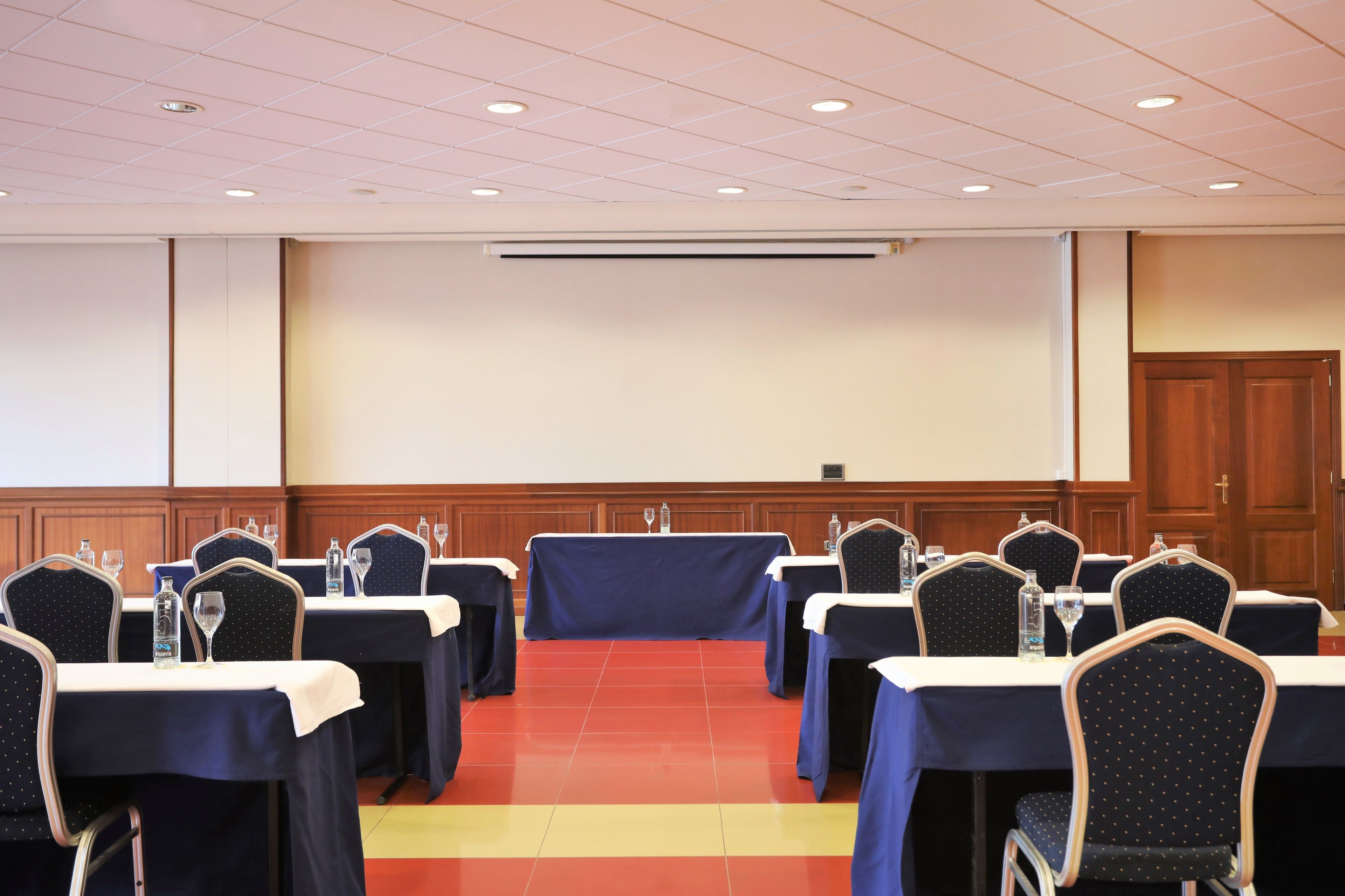 A conference room with tables and chairs, a projector screen, and a wooden panel wall.