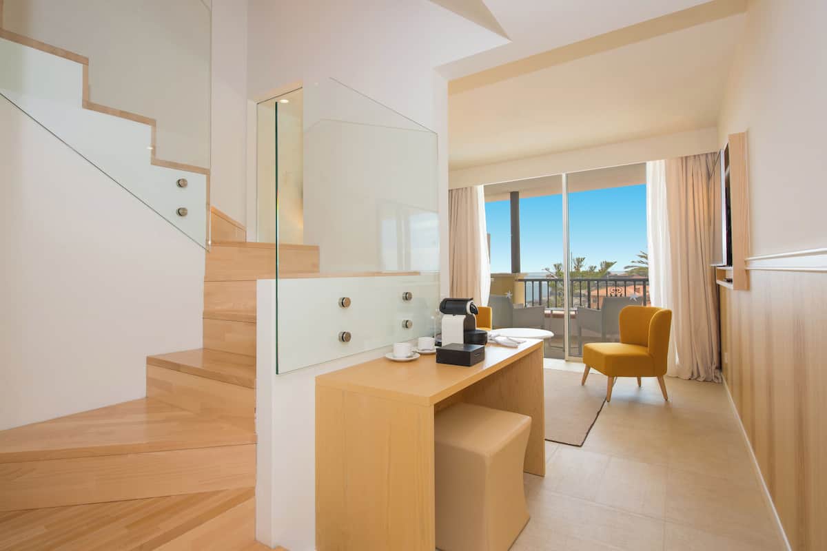 Quarto de hotel moderno com escada de madeira, secretária em vidro e uma poltrona amarela, além de varanda com vista.