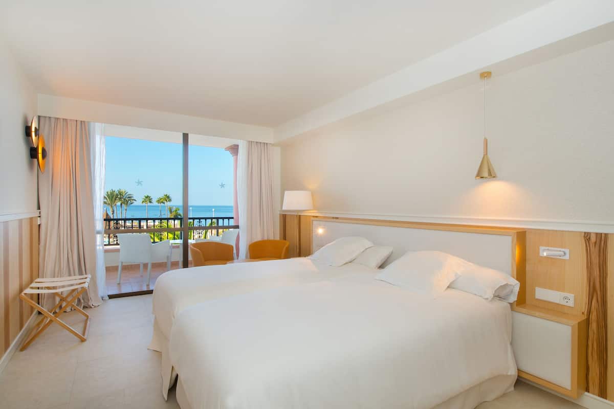Quarto de hotel com uma cama grande, varanda com cadeiras e vista para o mar.
