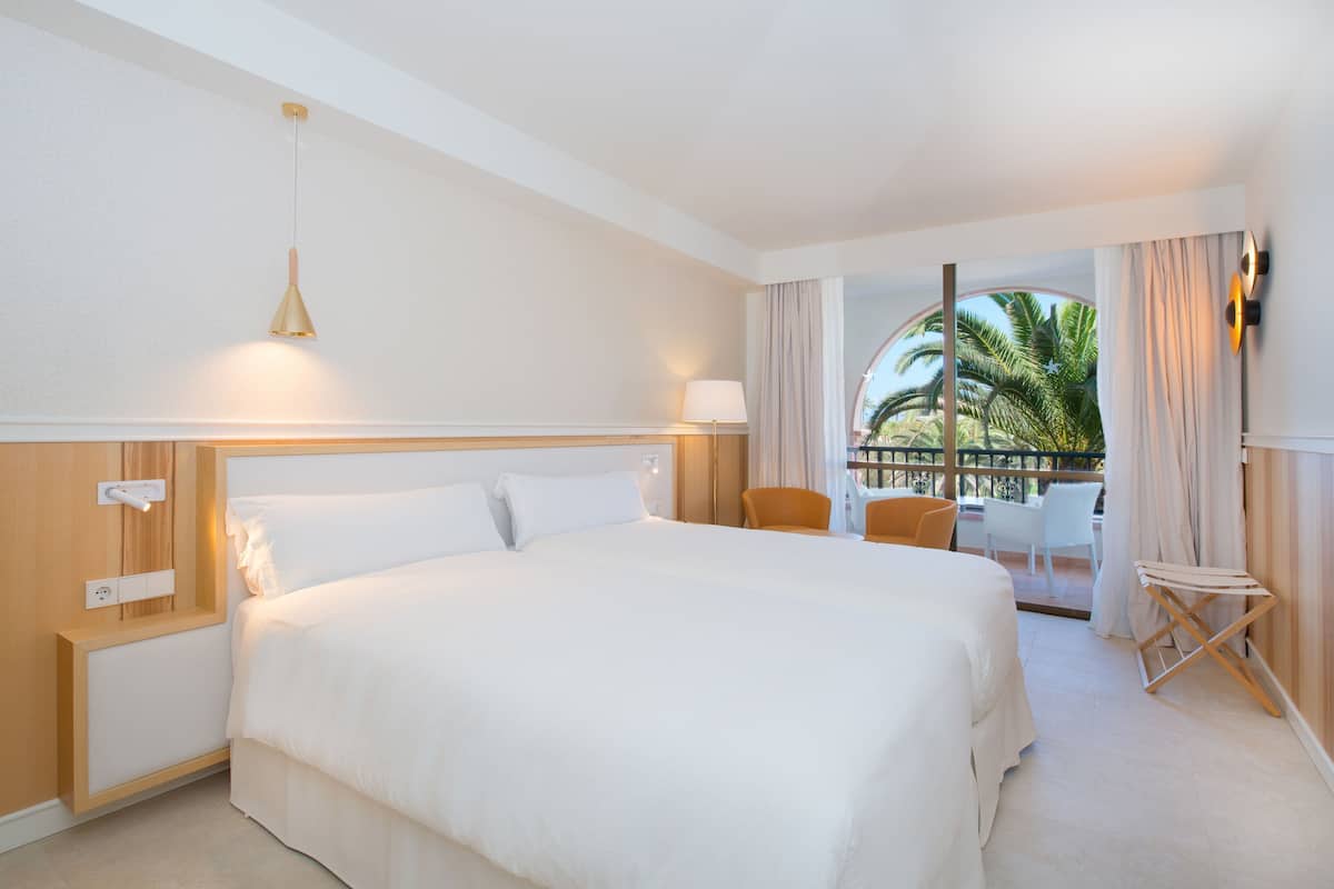 Quarto de hotel com uma cama grande, mesa de cabeceira, luminária, cadeira e vista para palmeiras.