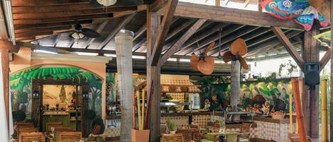 Un restaurante interior con vigas de madera, manteles verdes y un mural tropical.