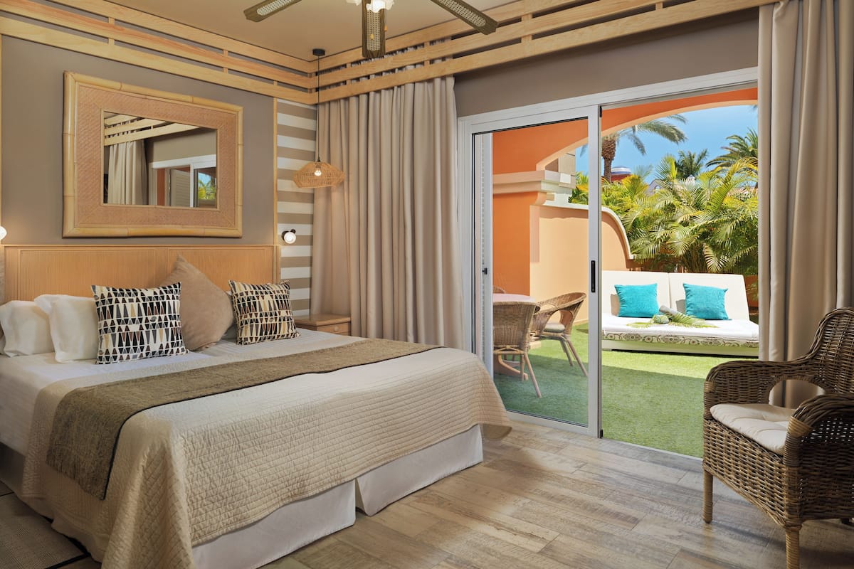 Ein Hotelzimmer mit einem großen Bett, einem Deckenventilator, einem Balkon mit Korbmöbeln und Blick auf Palmen.