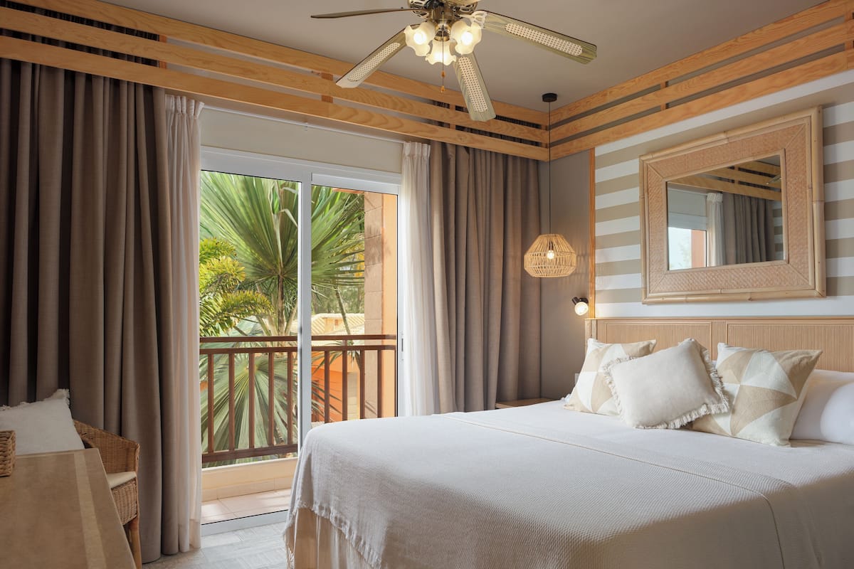 Ein Hotelzimmer mit Bett, Vorhängen, Deckenventilator, einem Fenster mit Blick auf Palmen und einem Spiegel an der Wand.