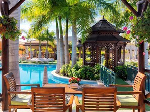 A poolside area with wooden chairs and a table, surrounded by palm trees and a gazebo. - GREEN GARDEN ECO RESORT & VILLAS (Arona)