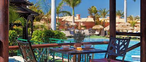 A poolside area with a table and chairs, a hanging planter, and a view of the pool and palm trees.