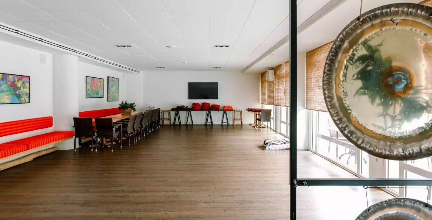 A spacious room with a long wooden table, red benches, and a large window.
