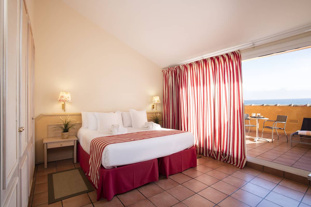 Ein Hotelzimmer mit Bett, Nachttischen, einer Topfpflanze, einem Balkon mit Meerblick und einem Tisch mit Stühlen.