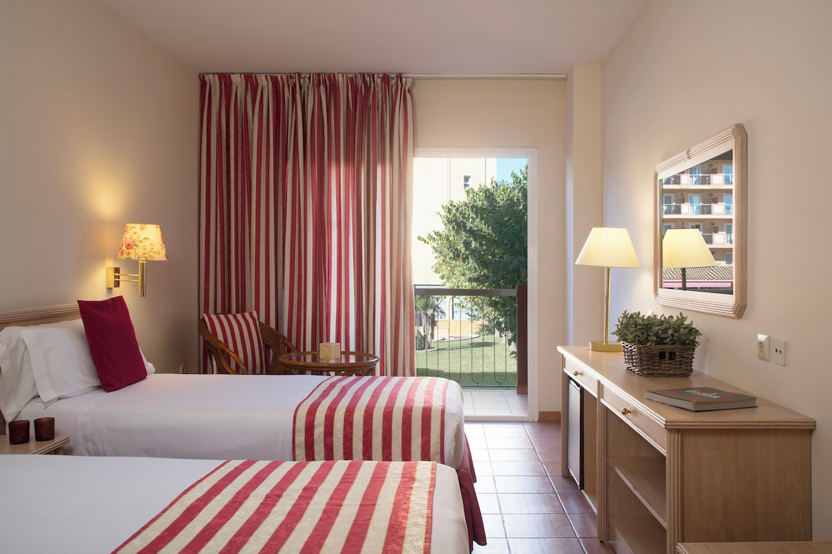 Ein Hotelzimmer mit zwei Betten, einem Schreibtisch, einem Stuhl, einem Balkon mit Blick und einem fenster mit Vorhängen.