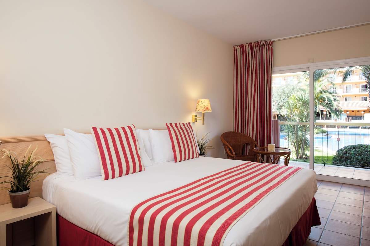 Une chambre d’hôtel avec un grand lit, une petite table, une chaise, une plante en pot et une vue sur une piscine et les bâtiments environnants.