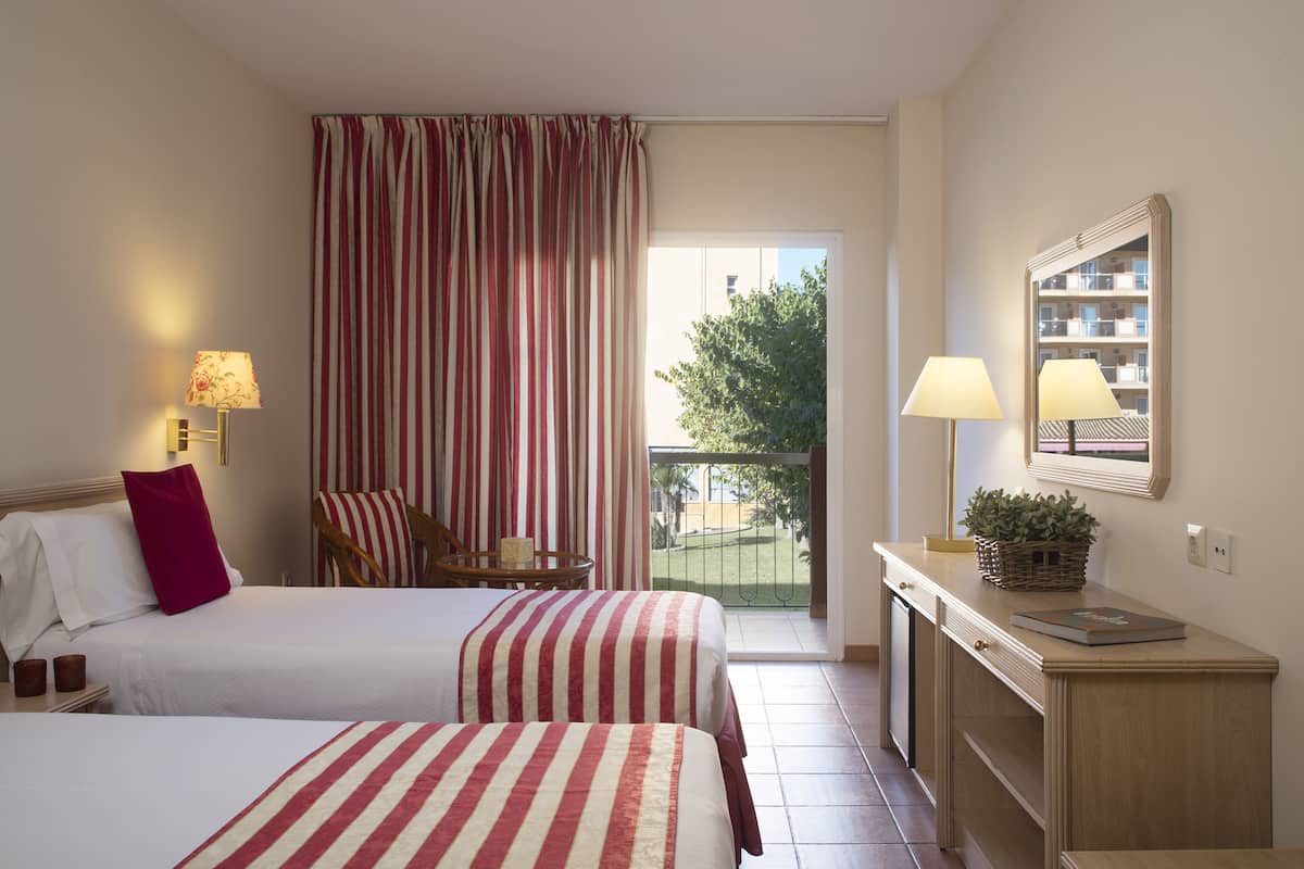 Ein Hotelzimmer mit zwei Betten, einem Schreibtisch, einem Stuhl, einem Balkon mit Blick und einem fenster mit Vorhängen.