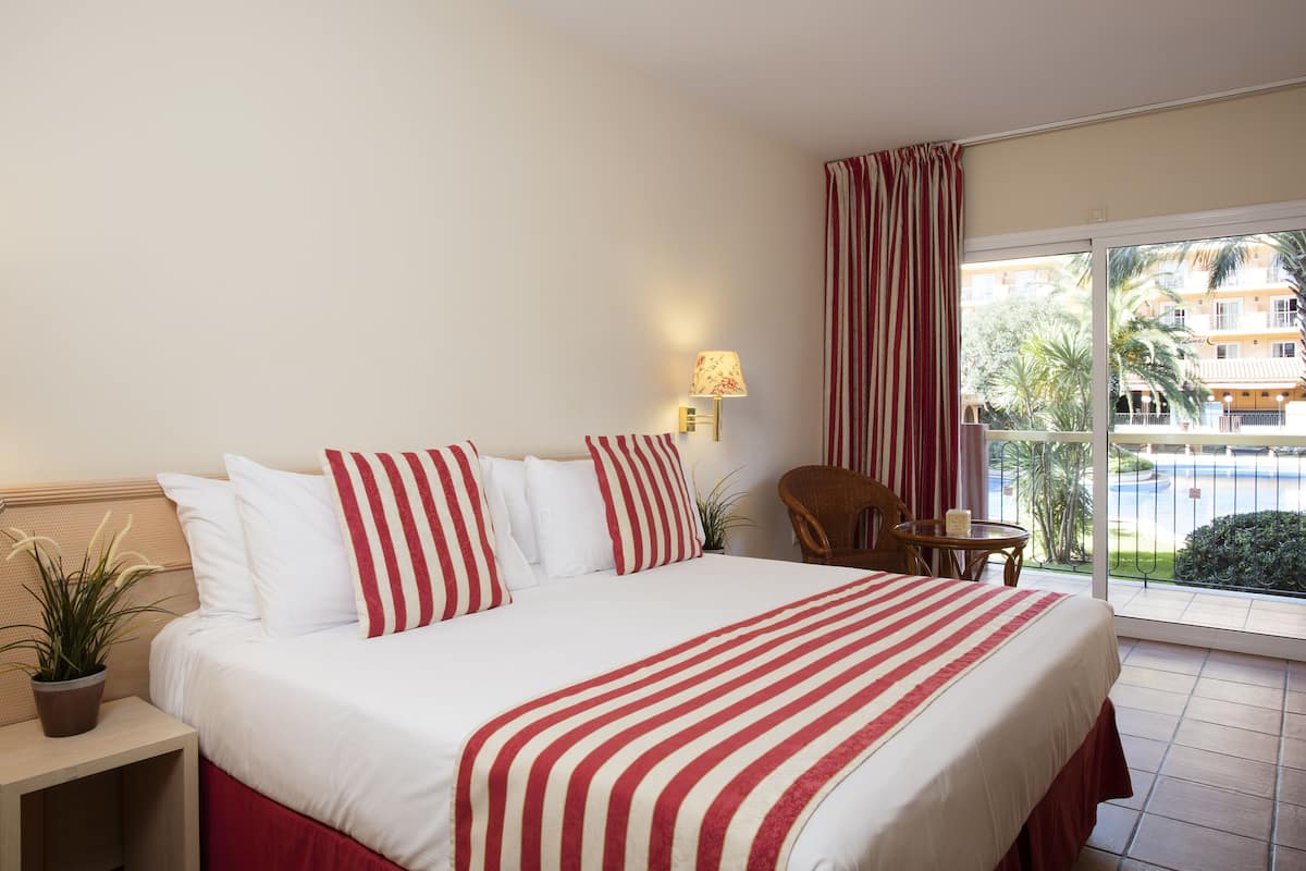 Ein Hotelzimmer mit einem großen Bett, einem kleinen Tisch, einem Sessel, einer Topfpflanze und Blick auf einen Pool und Gebäude.