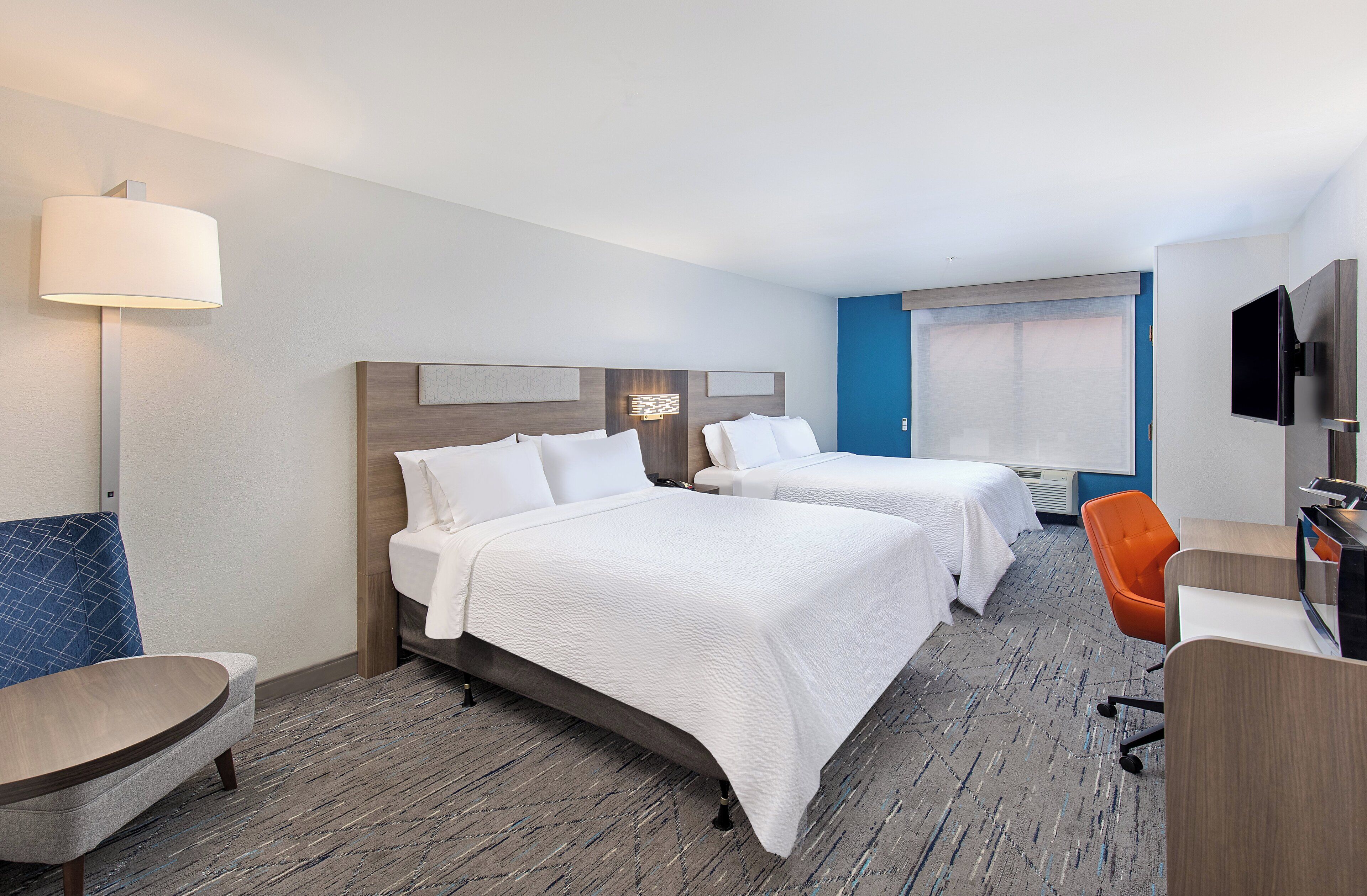 A hotel room with two beds, a desk, a chair, a TV, and a lamp.