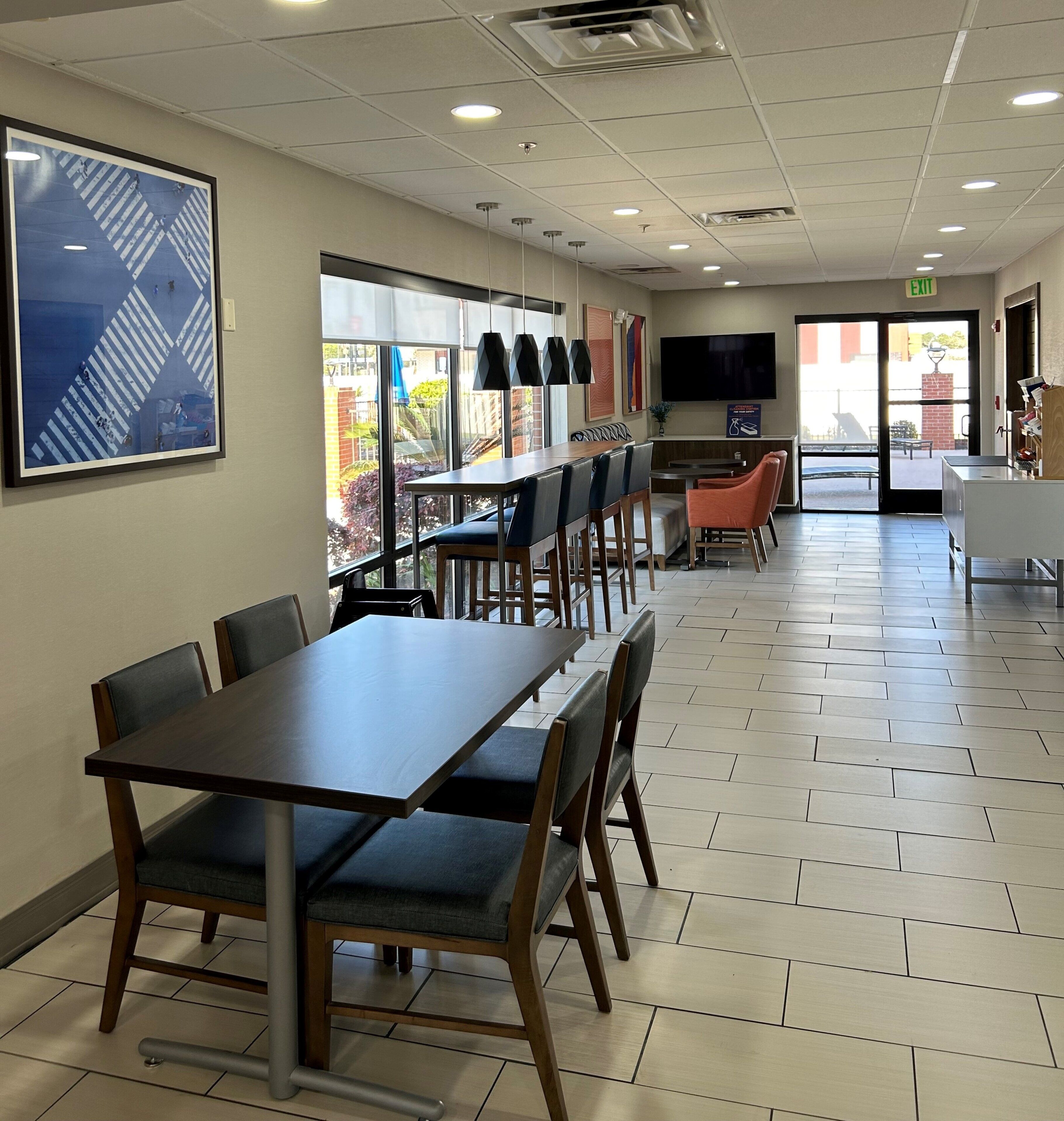 A spacious dining area with multiple tables and chairs, a large window with a view of the outdoors, and a flat-screen TV mounted on the wall.