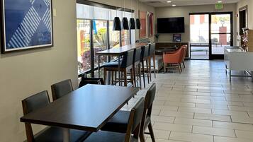 A spacious dining area with multiple tables and chairs, a large window with a view of the outdoors, and a flat-screen TV mounted on the wall.