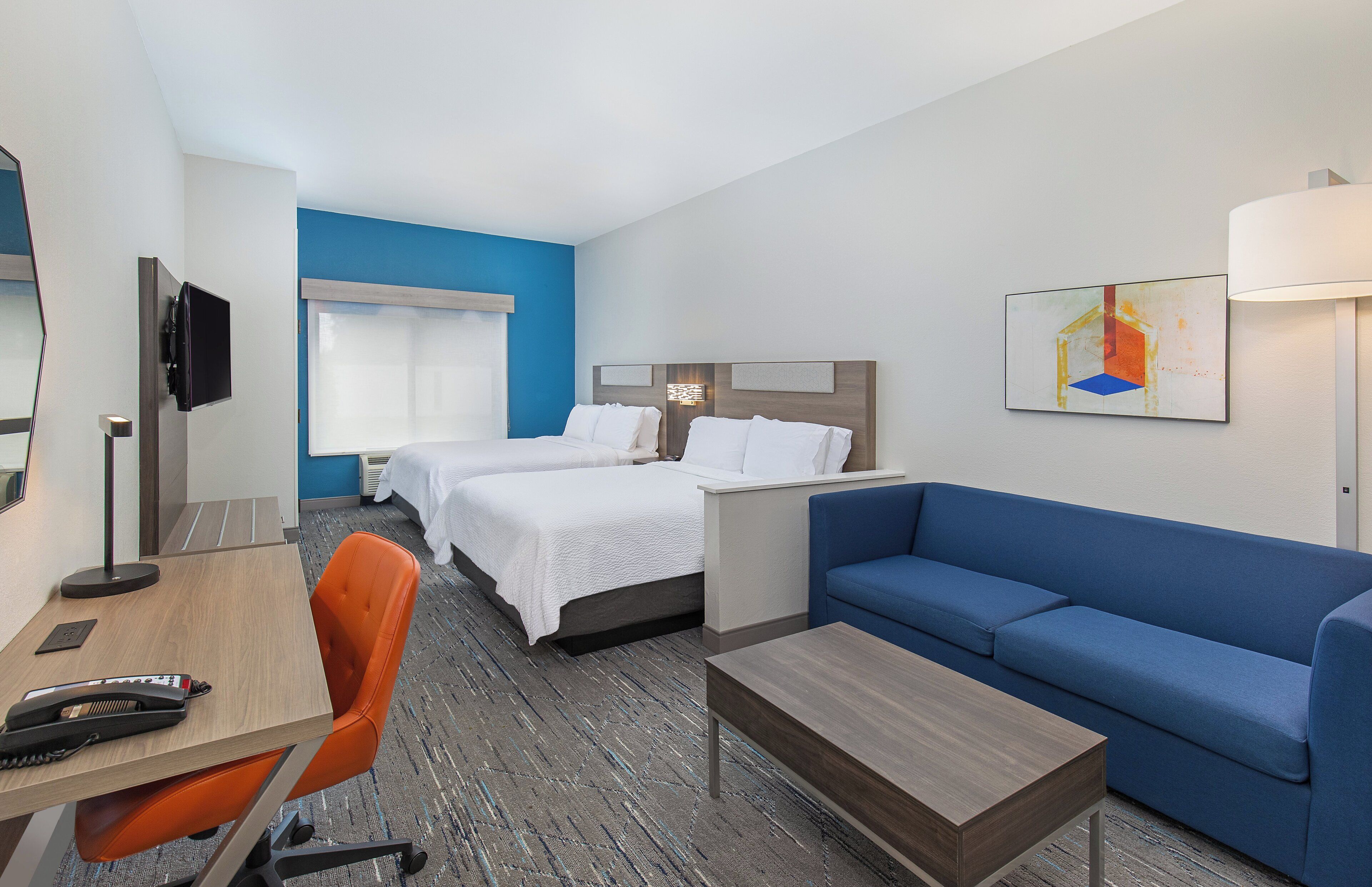 Quarto de hotel com duas camas, uma escrivaninha, uma cadeira, um sofá e uma televisão.