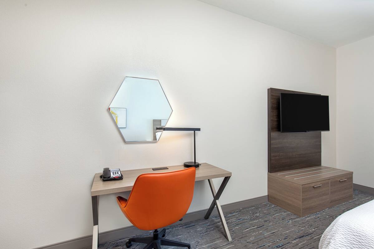 Une chambre d’hôtel équipée d’un bureau, d’un fauteuil orange, d’un miroir, d’un téléviseur à écran plat et d’une armoire en bois.