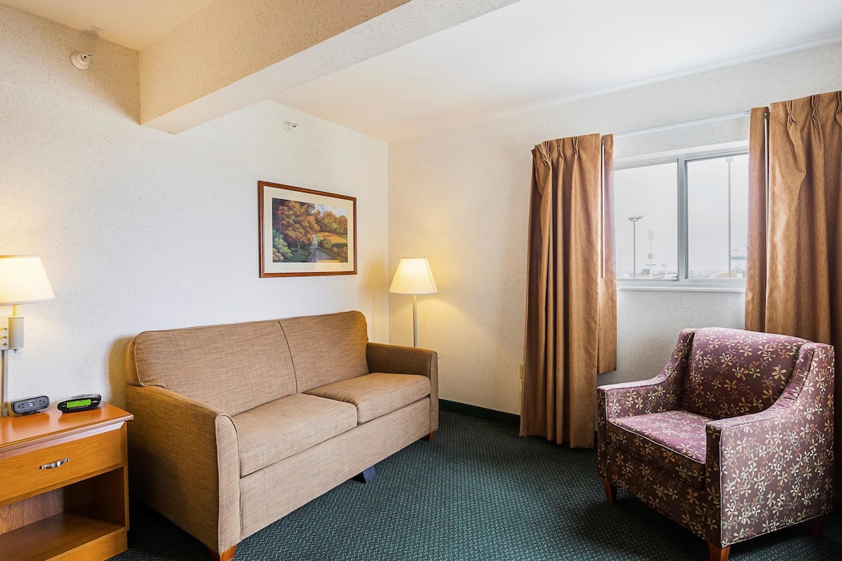 Habitación de hotel con sofá, sillón, mesita de noche, lámparas y ventana con cortinas.