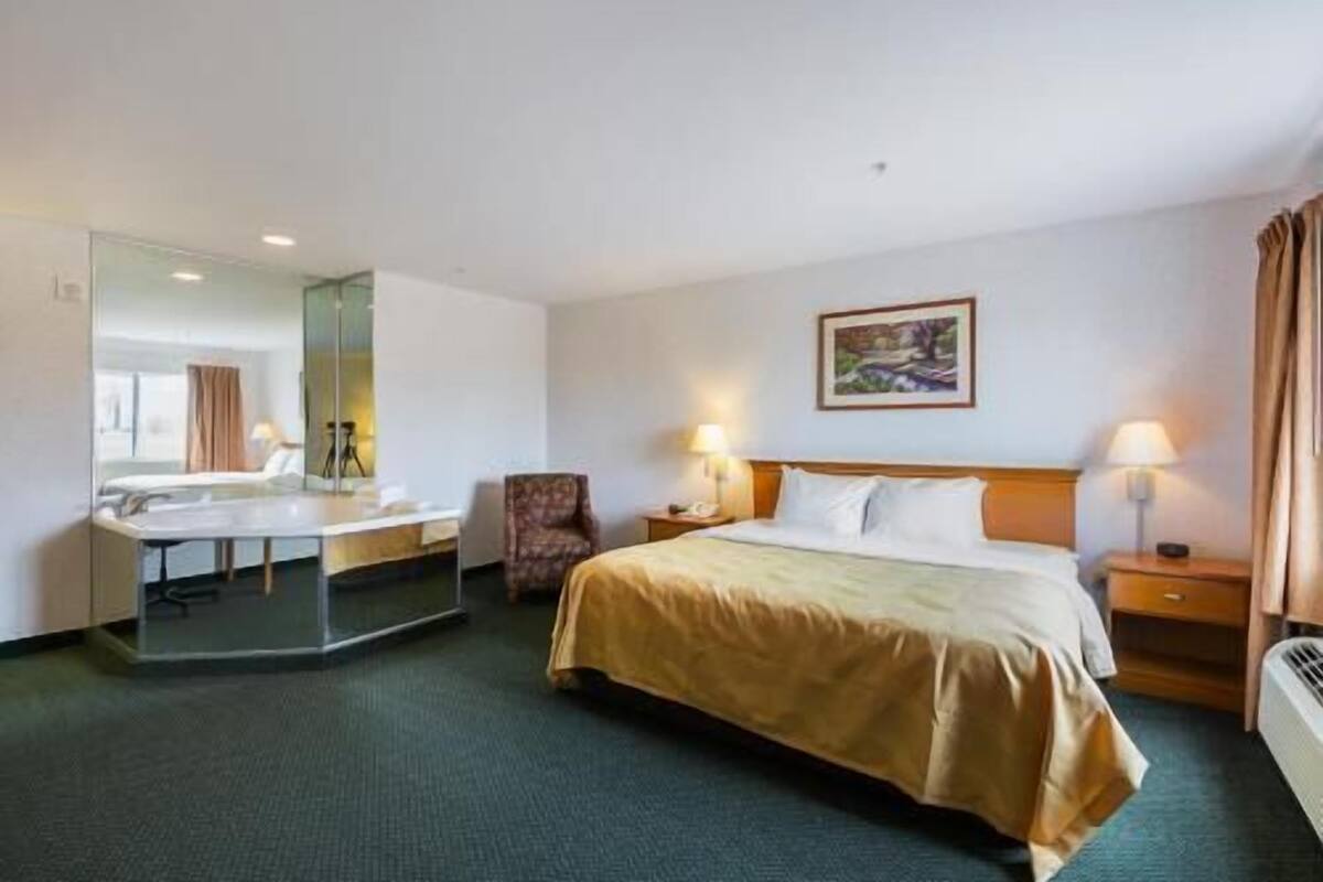 Una habitación de hotel con una cama, un escritorio con silla, un espejo, una mesita de noche con lámpara y un cuadro en la pared.
