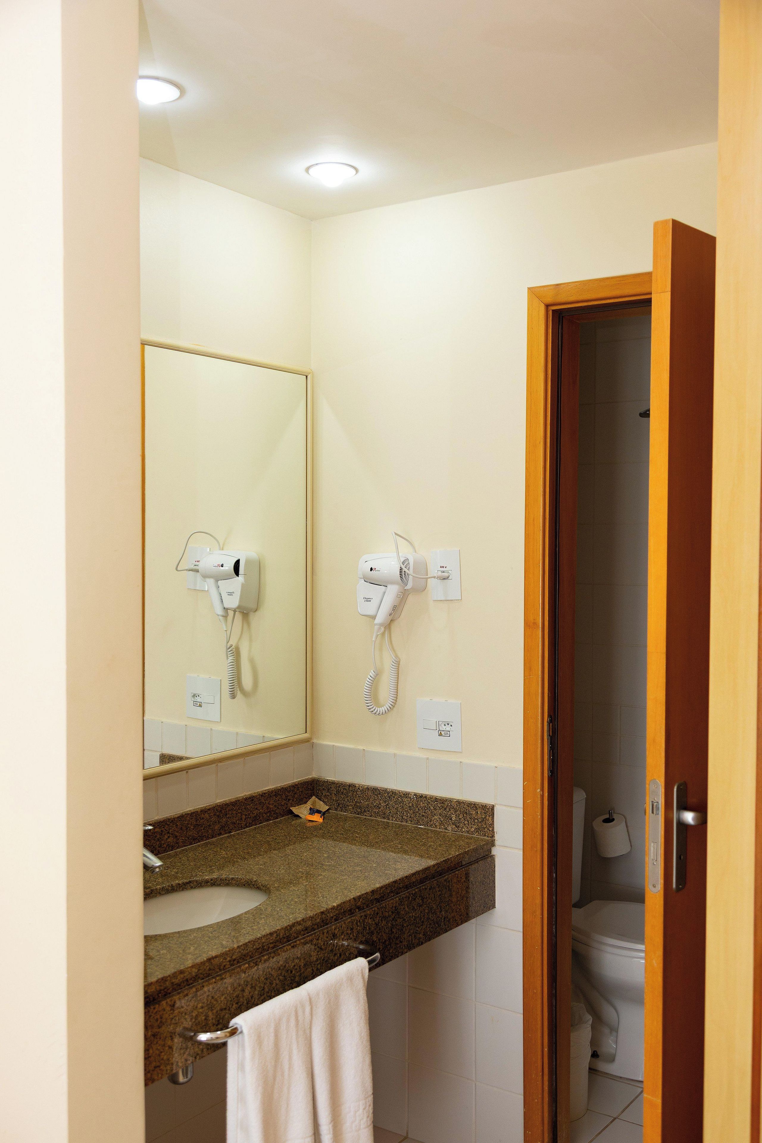 A bathroom with a granite countertop, two hair dryers mounted on the wall, a mirror, and a toilet visible through an open door.