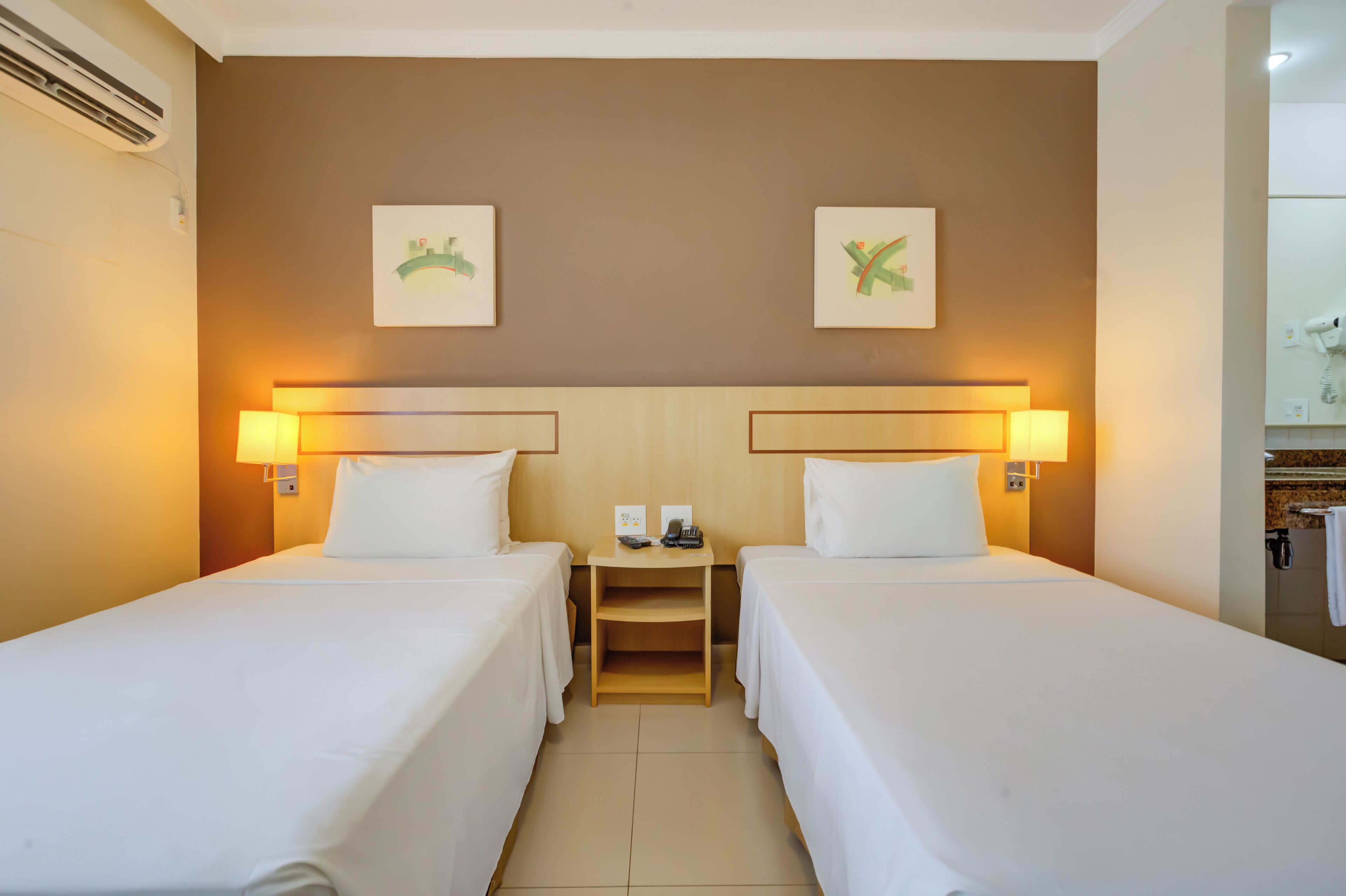 A hotel room with two single beds, a wooden nightstand with a phone and a lamp, and a bathroom visible to the right.