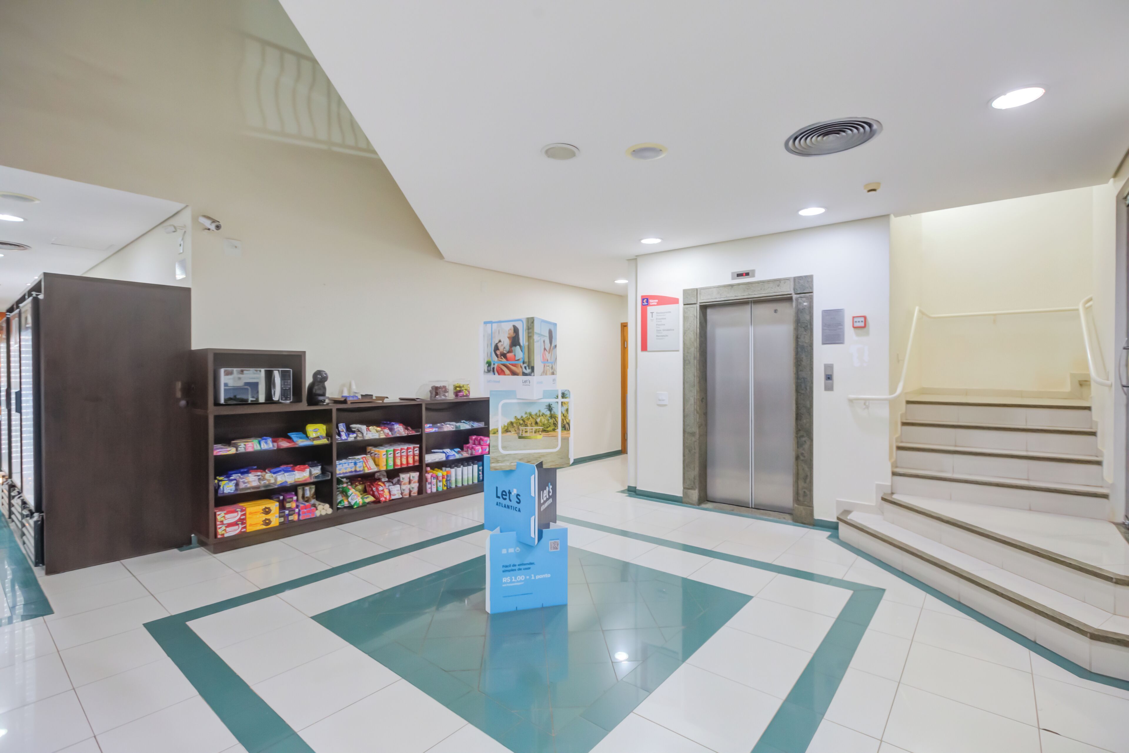 Eine Lobby mit Empfangsschalter, Getränkeautomaten, einer Treppe und einem digitalen Aufzugsanzeigebildschirm.