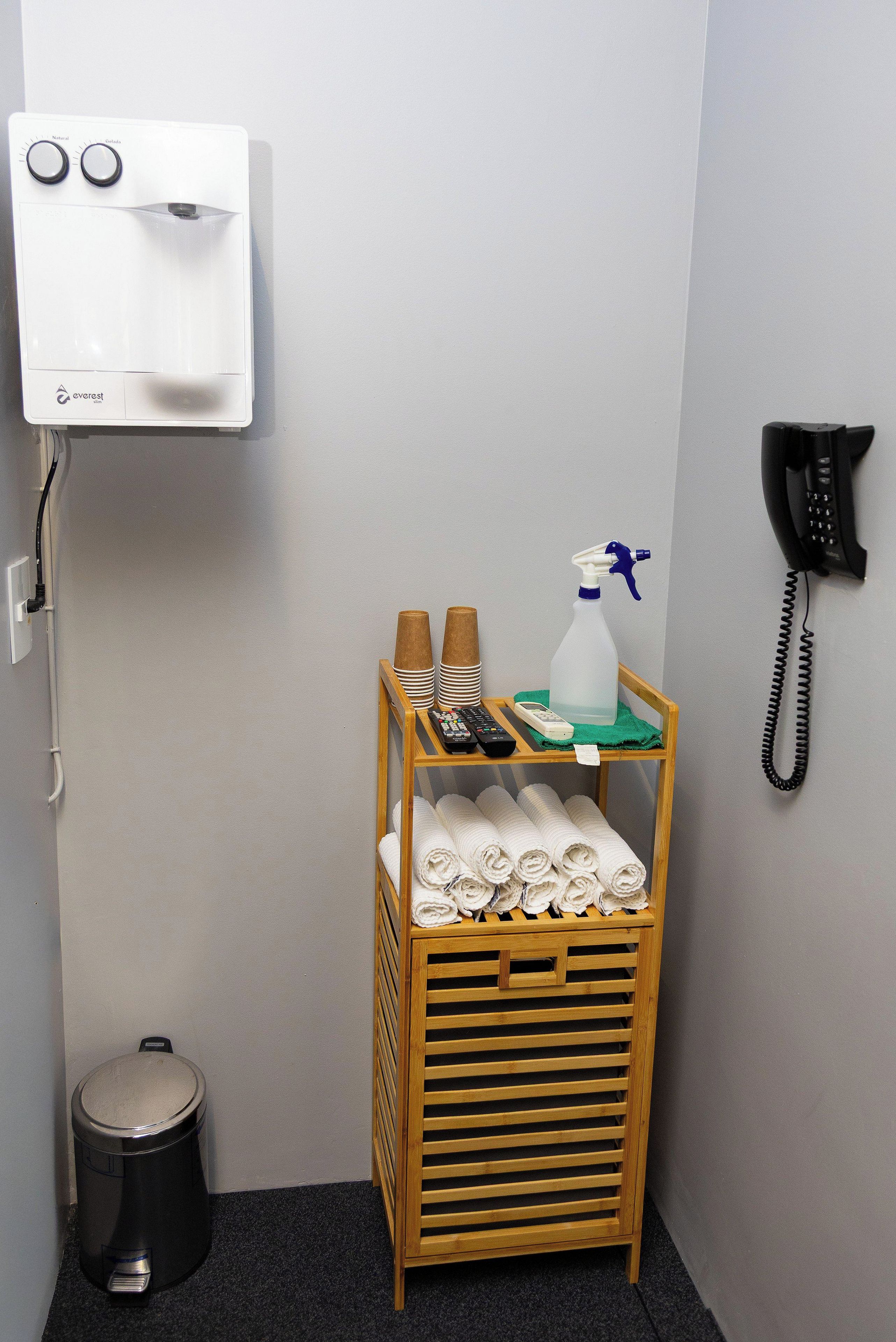 A small hotel room with a white wall, a wooden shelf with towels, a spray bottle, and a telephone.