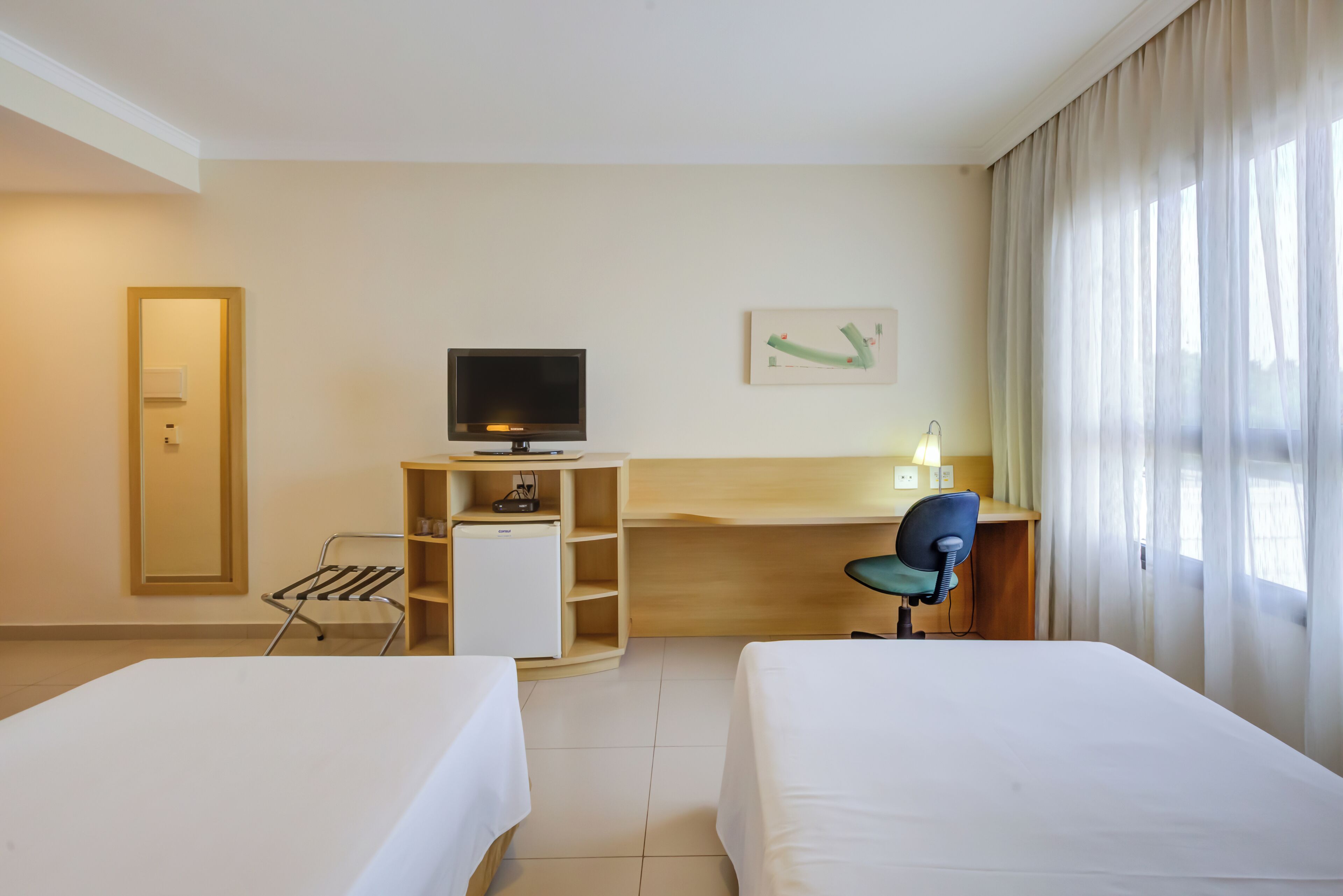A hotel room with two beds, a desk with a TV, a chair, and a window with curtains.