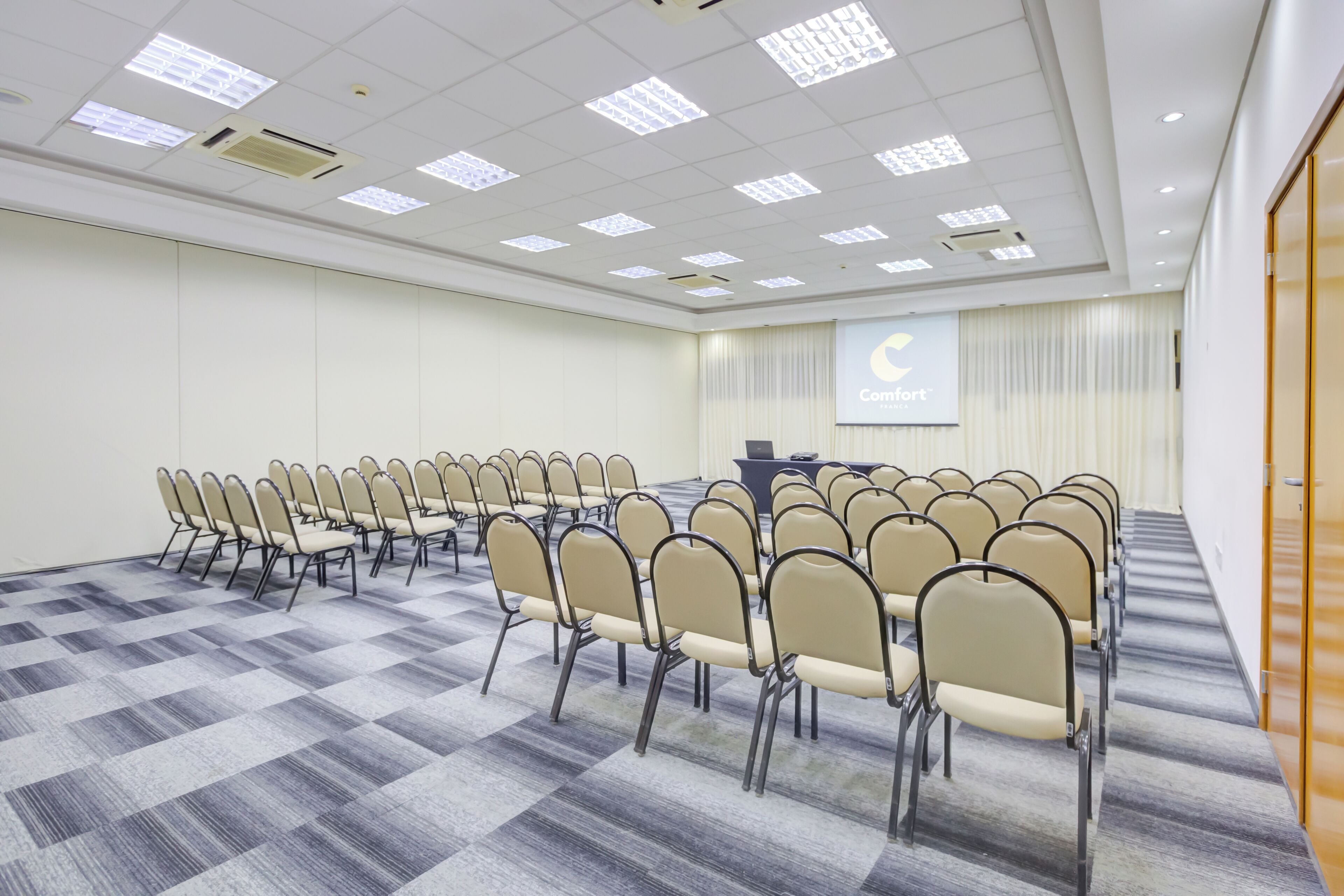 Une salle de conférence avec des rangées de chaises, un pupitre et un écran affichant le logo de Comfort.