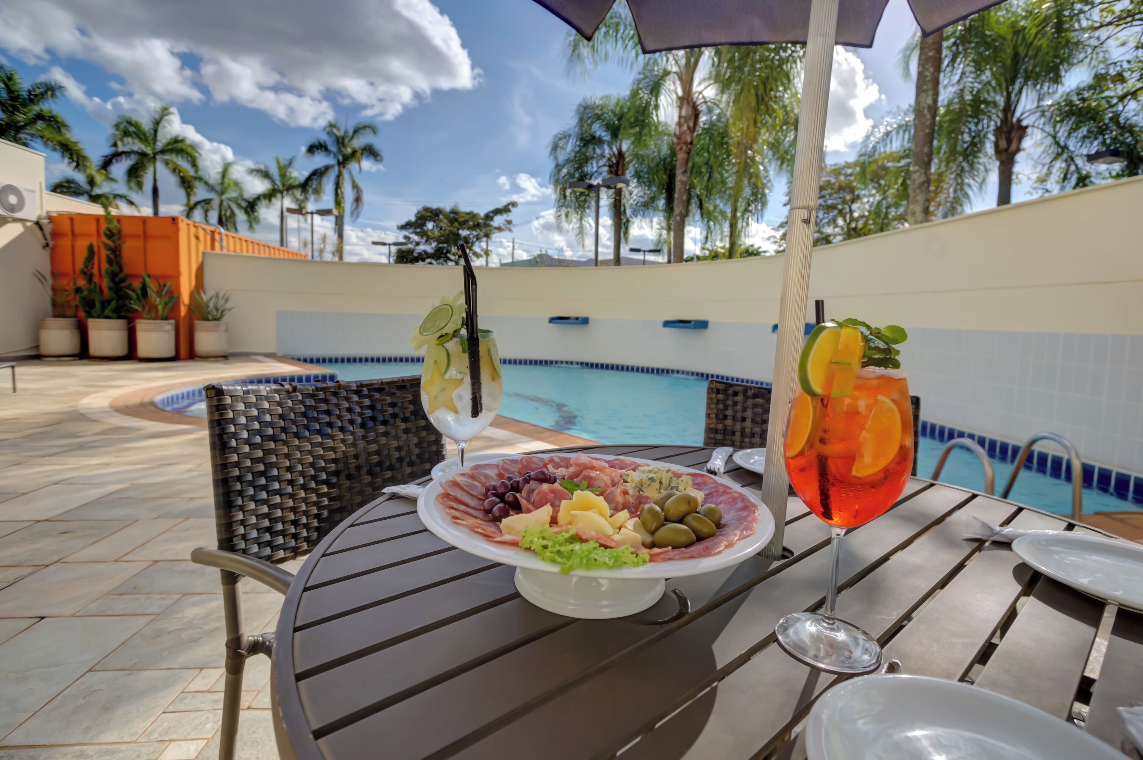Un espace extérieur aménagé pour les repas, avec une piscine, une table garnie d’un assortiment de fruits et de boissons, et une vue sur des palmiers.