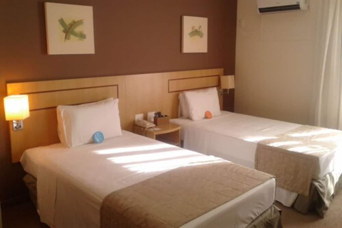 Una habitación de hotel con dos camas individuales, una mesita de noche con lámpara, un teléfono y una pequeña mesa con un libro.