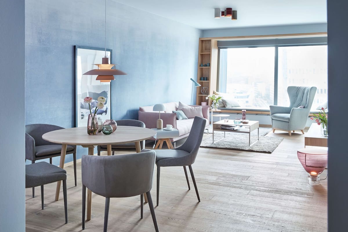 A modern living room with a round table, grey chairs, a sofa, and a large window offering a city view.