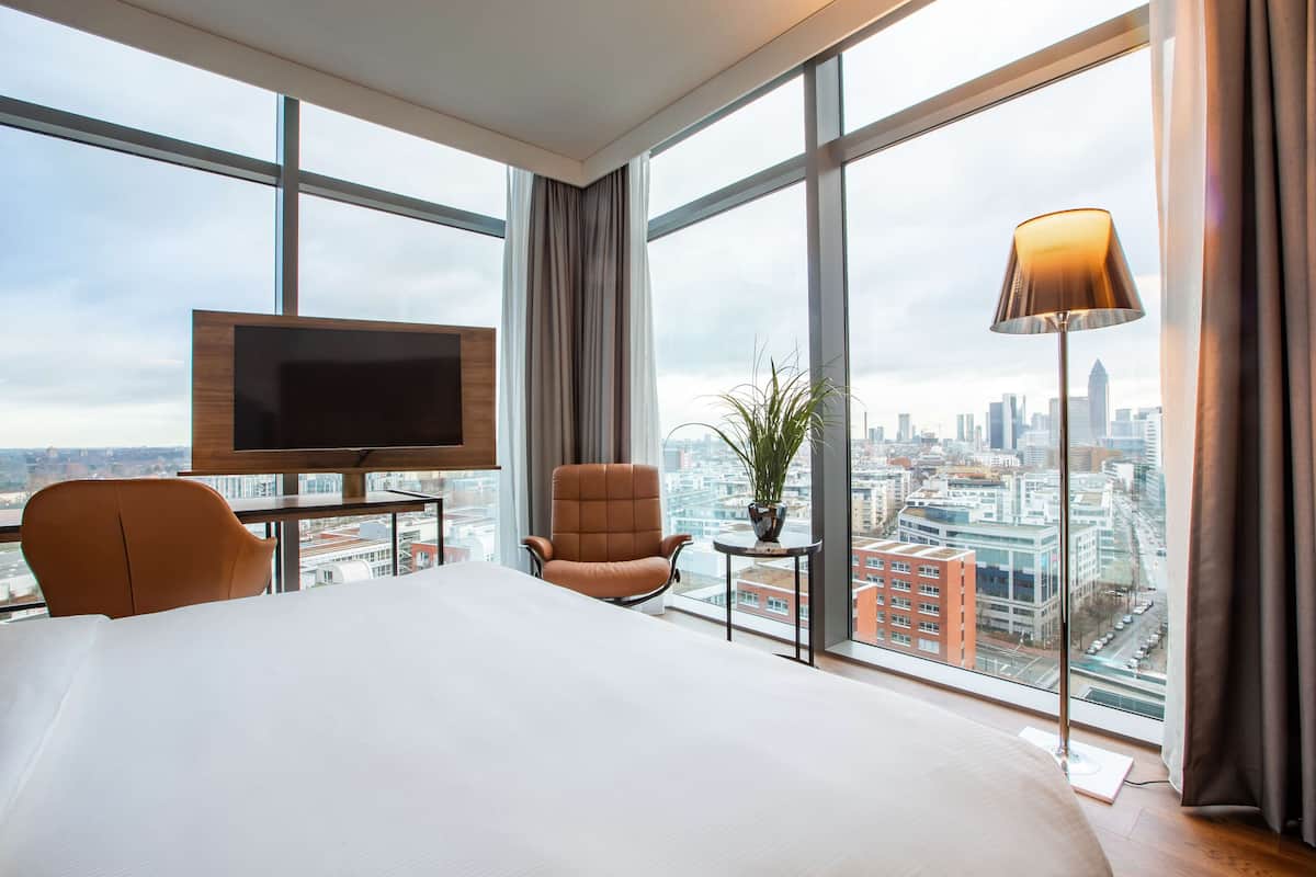 A hotel room with a large bed, a TV, a chair, a small table with a plant, and a view of a cityscape through large windows.