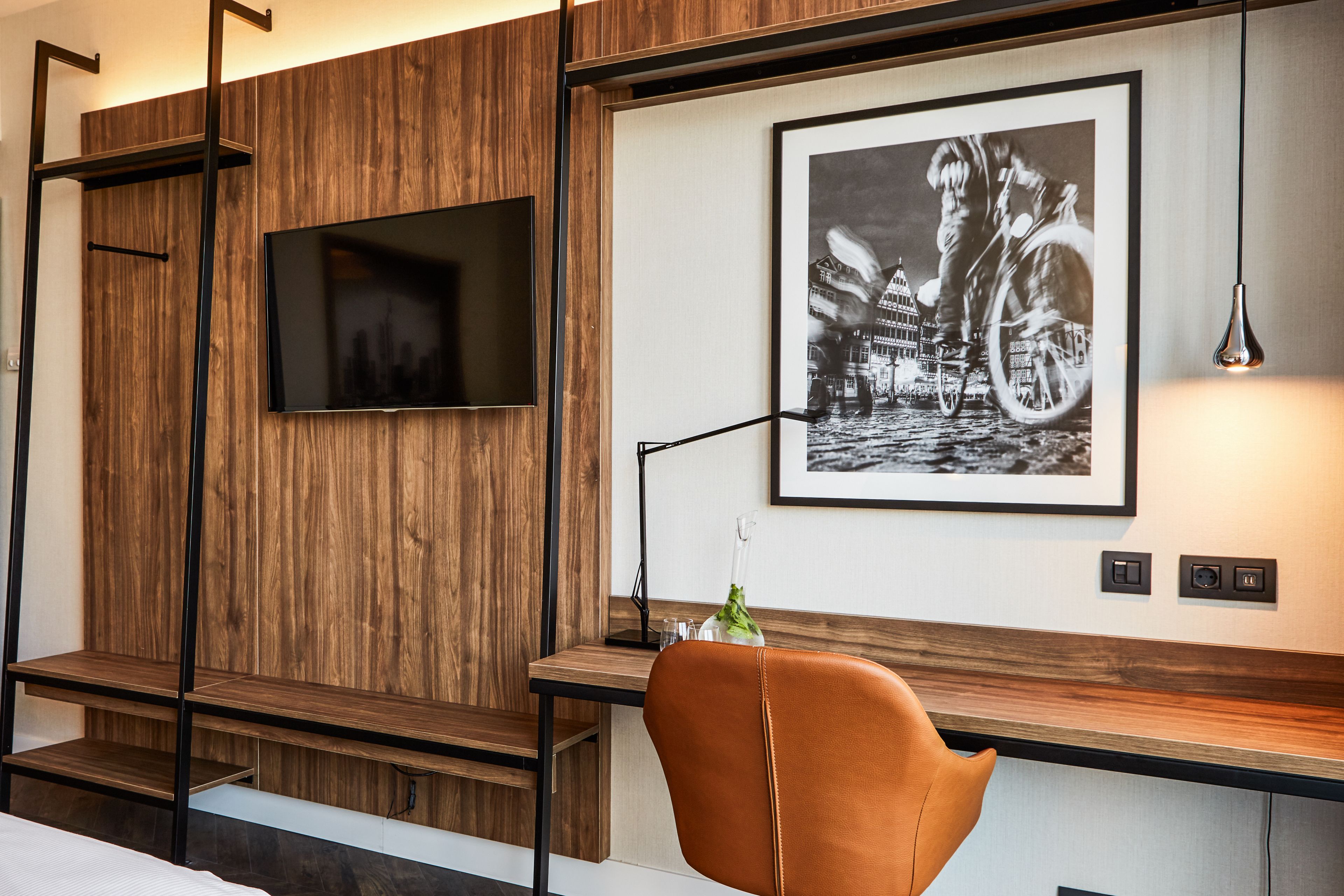 Une chambre moderne avec un mur en bois, un téléviseur fixé au mur, une photographie en noir et blanc encadrée et un fauteuil marron.
