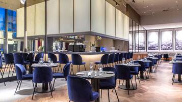 Un restaurante moderno con mesas redondas y sillas azules, una zona de bar y grandes ventanales que ofrecen vistas a la ciudad.
