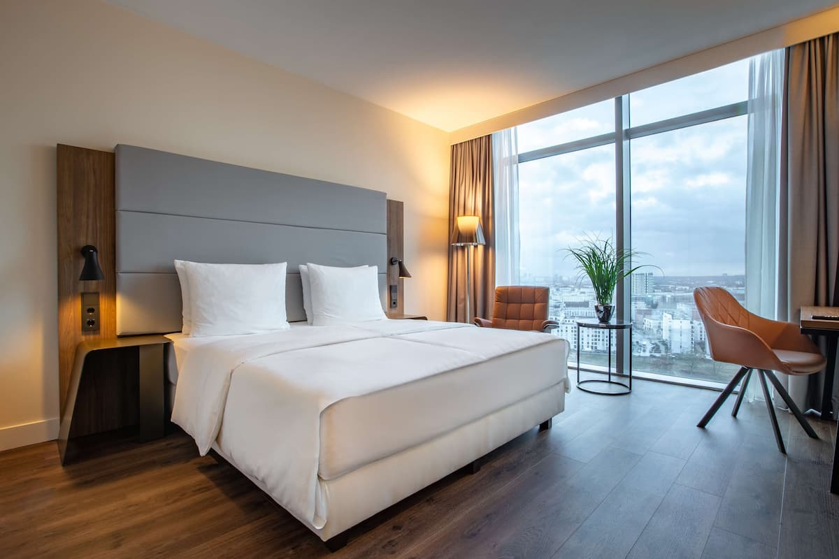 A modern hotel room with a large bed, a sitting area with a chair, a small table, and a large window offering a city view.