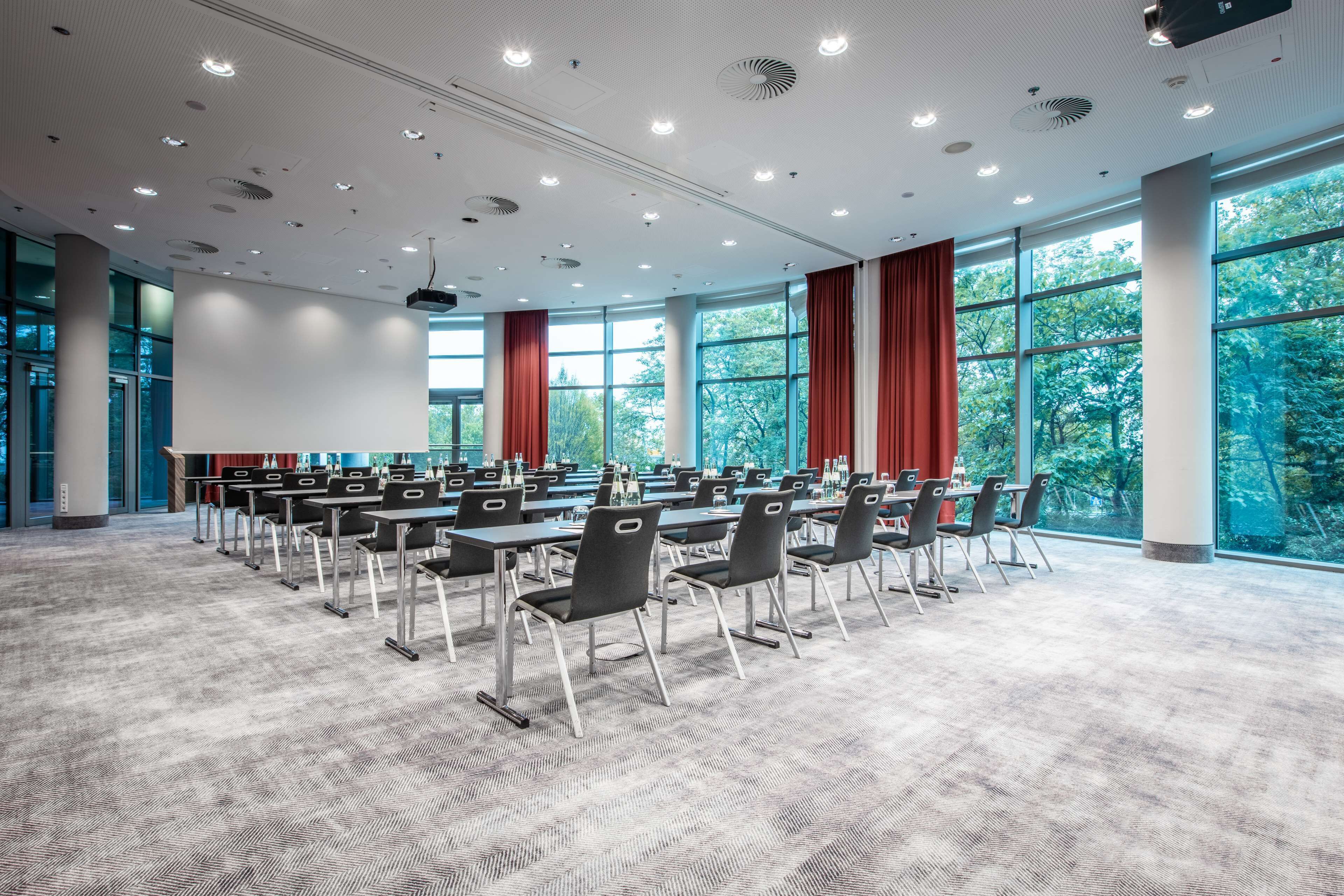 A conference room with rows of chairs and tables, large windows with red curtains, and a carpeted floor.