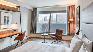 A hotel room with a large bed, a desk, two chairs, a small table with a glass, and a view of the city skyline through a large window. - Radisson Blu Hotel, Frankfurt (Frankfurt)