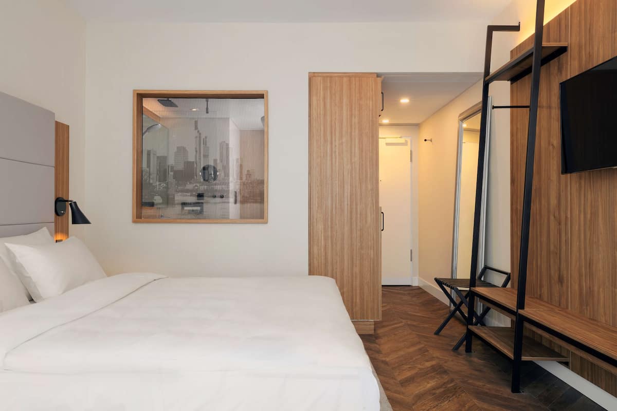 A modern hotel room with a large bed, a wooden wardrobe, a TV mounted on the wall, and a cityscape view through a window.