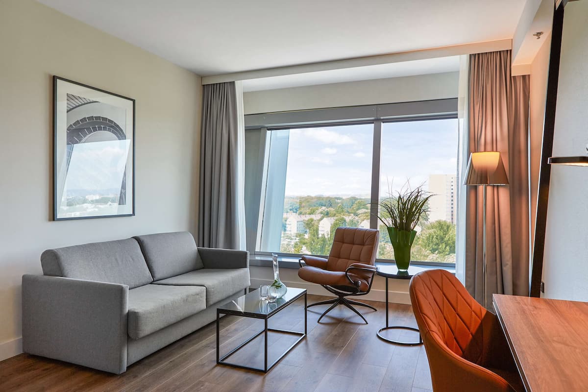 A modern living room with a grey sofa, a wooden coffee table, an orange armchair, and a view of a cityscape through large windows.
