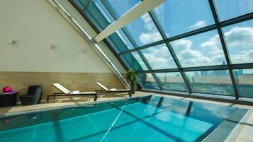 An indoor swimming pool with clear glass walls, lounge chairs, and a potted plant.