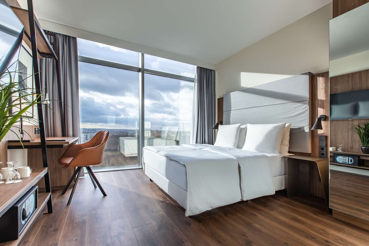 A modern hotel room with a large bed, a desk with a chair, a large window with curtains, and a view of the city.