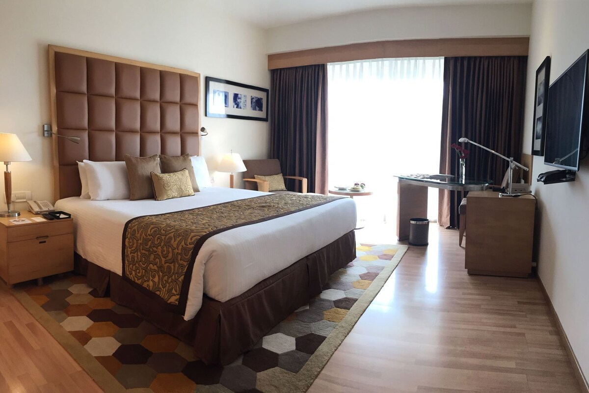 A hotel room with a large bed, bedside tables, a desk, a chair, a TV, and a window with curtains.