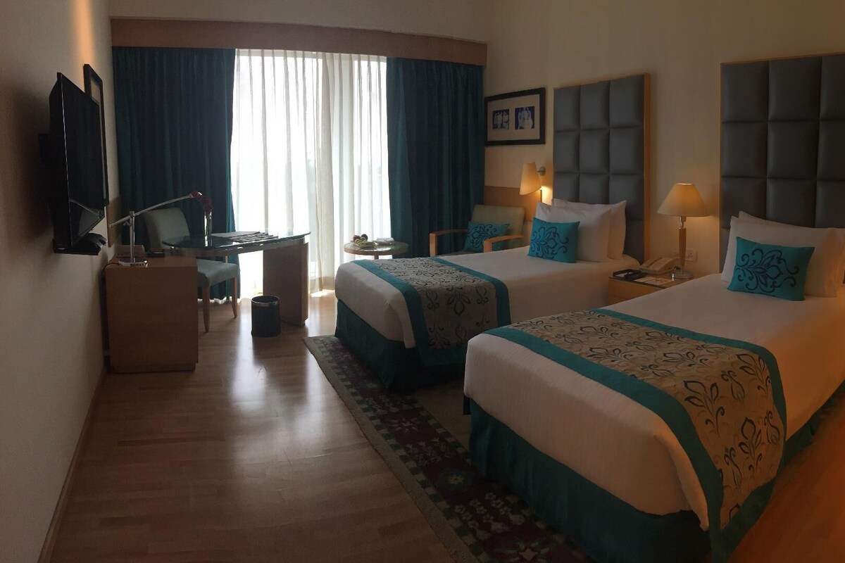 A hotel room with two beds, a desk, a chair, a TV, and a window with curtains.