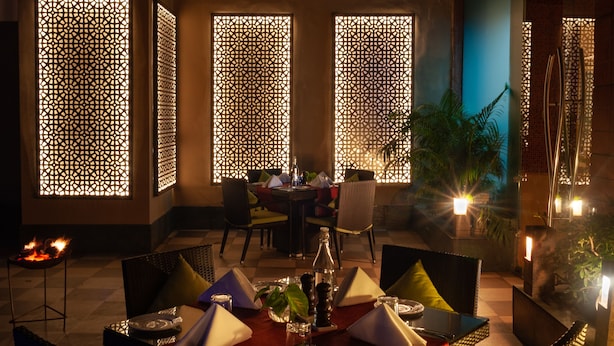 Un coin repas avec une table dressée pour deux personnes, entouré de chaises en osier agrémentées de coussins. Les murs sont ornés de paravents décoratifs aux motifs géométriques, et un petit foyer se trouve sur le côté gauche.